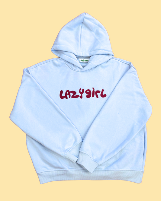 A light blue pullover hoodie with 'LAZY girl' printed in red across the chest, laid flat on a pale yellow background.
