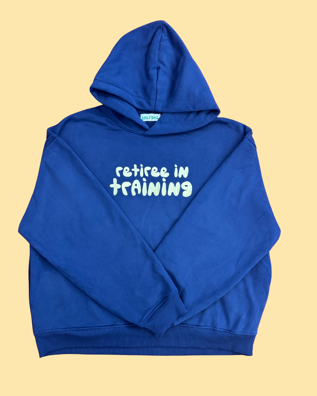 A royal blue pullover hoodie with the phrase 'retiree in training' printed in pale yellow across the chest, laid flat on a solid pale yellow background.