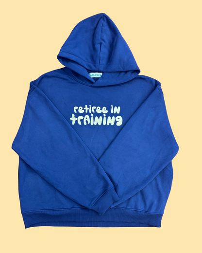 A royal blue pullover hoodie with the phrase 'retiree in training' printed in pale yellow across the chest, laid flat on a solid pale yellow background.