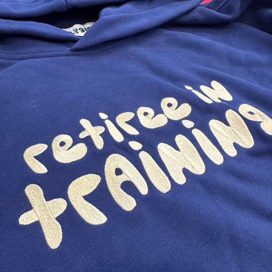 A close-up of a navy blue hoodie with the phrase 'retiree in training' printed across the chest in a light beige bubble font.